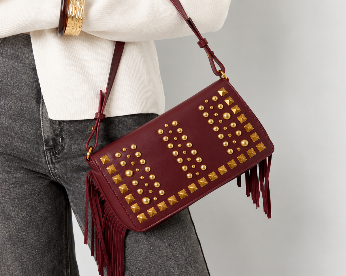 Model met bordeauxrode schoudertas met goudkleurige studs en franjes, gedragen bij een lichte trui en donkere jeans.